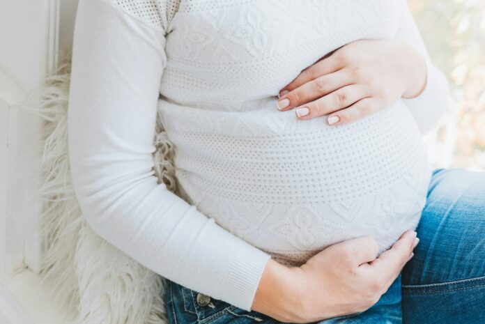 Pregnant woman in a white sweater and jeans gently holding her baby bump while sitting by a window.
