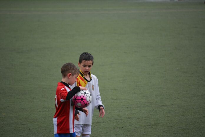 Youth football players talking on the pitch during a match, with one holding a ball.