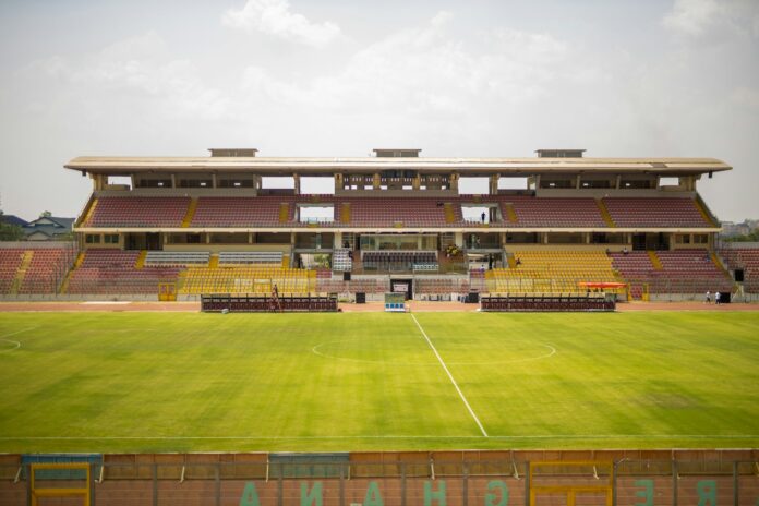 View of Baba Yara Stadium in Kumasi, Ghana, showing an empty pitch and stands with red, yellow, and white seating under a clear sky.