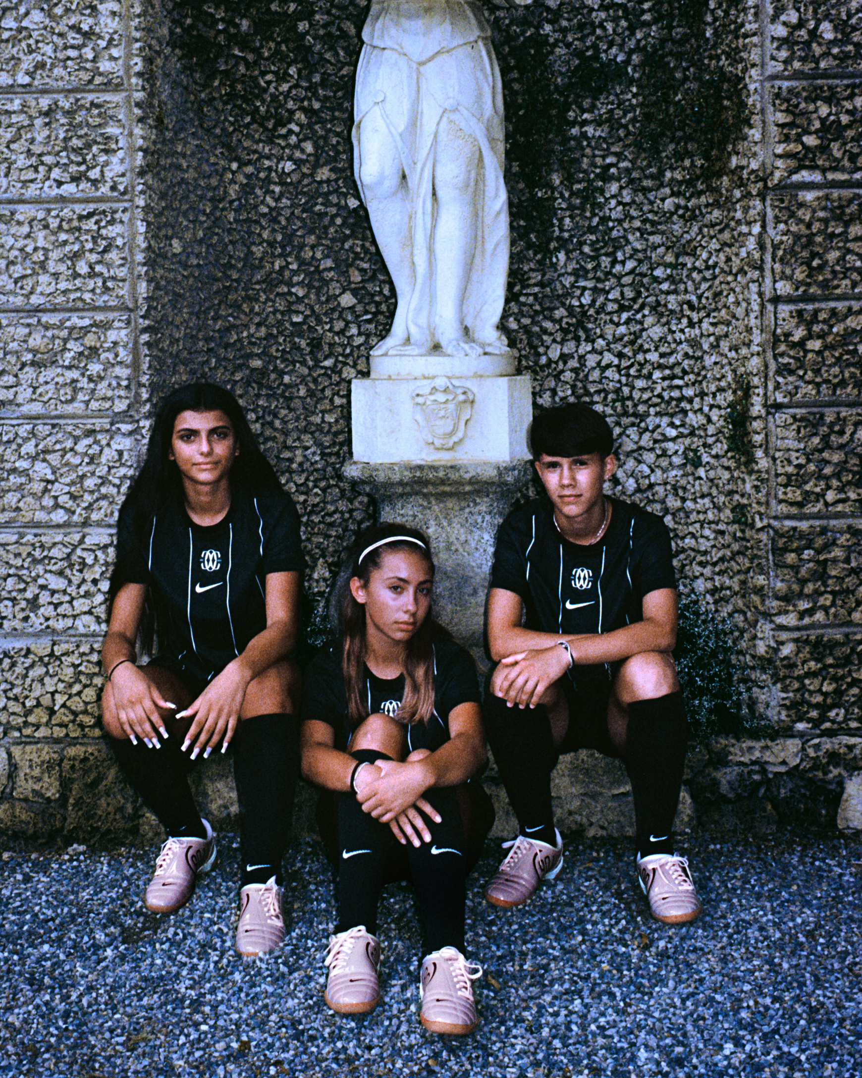 Three F.C. Como Women youth players wearing the 2025/26 Home kit sit in front of a stone statue during The Next Wave campaign shoot.