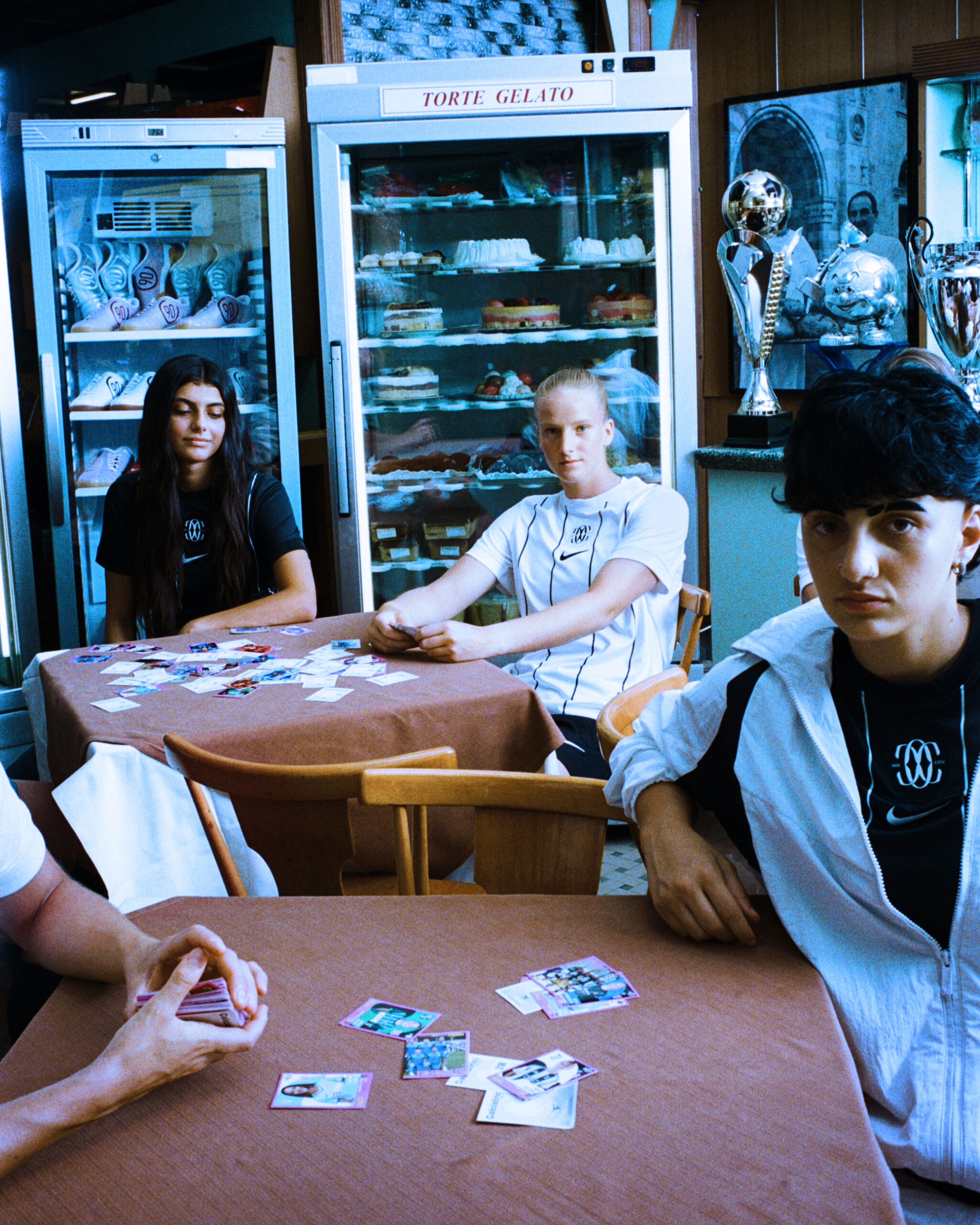 F.C. Como Women players sit at café tables covered with football stickers during The Next Wave campaign shoot, with trophies and desserts displayed in the background.