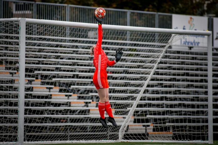 Goalkeeper in red kit leaps to tip the ball over the crossbar during training, highlighting agility and game-realistic decision-making