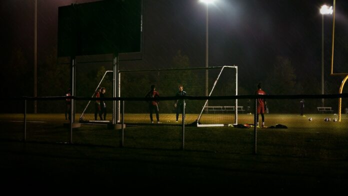 Goalkeepers and a coach train under floodlights on a rainy night, preparing near the goalpost in a dimly lit football field