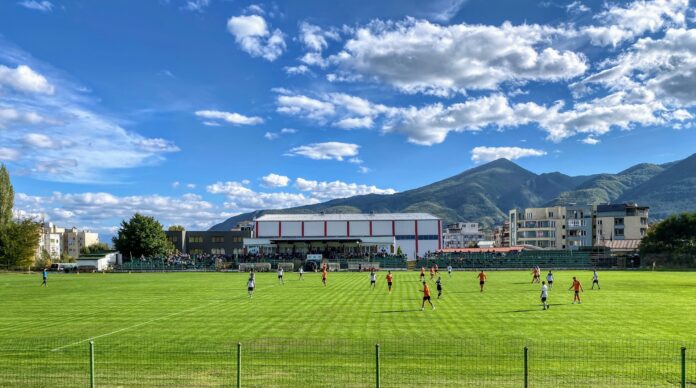 A local football match taking place on a green pitch in Gotse Delchev, Bulgaria, with mountains and residential buildings in the background.