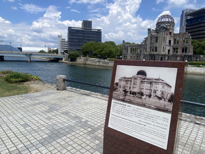 View of the Hiroshima Peace Memorial (Atomic Bomb Dome) with a historical information plaque in the foreground beside the Motoyasu River