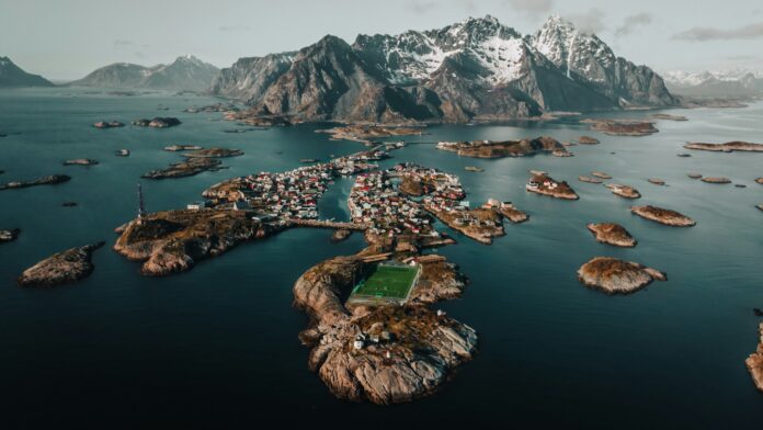 Aerial view of a coastal Scandinavian village surrounded by islands and mountains, featuring a football pitch prominently placed on a rocky outcrop.