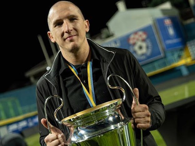 Toni Bilandžić holding a championship trophy on the football pitch, wearing a medal around his neck.