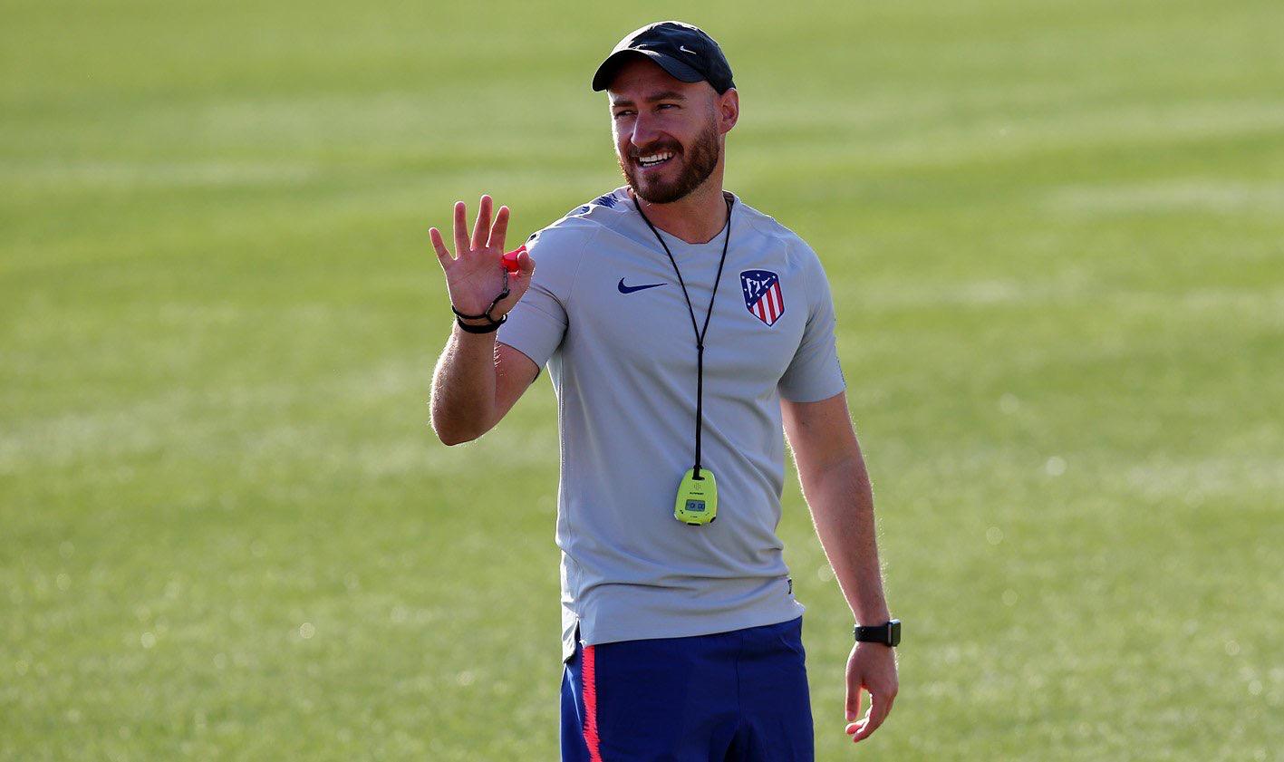 Carlos González smiles during an Atlético Madrid youth training session, wearing club gear and holding a whistle on the pitch.