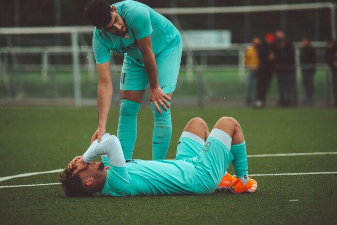 A football player lies on the pitch holding his head after a collision while a teammate checks on him, highlighting the risks of concussions in football.