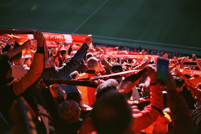 Liverpool fans holding scarves aloft at Anfield, symbolizing the global reach and commercial impact of celebrity owners like LeBron James.