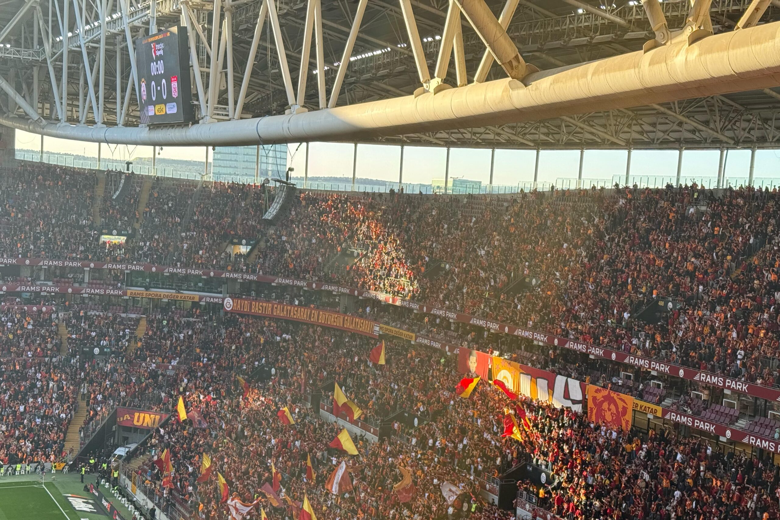 Galatasaray supporters waving flags and banners in the stands at Rams Park during a home match.