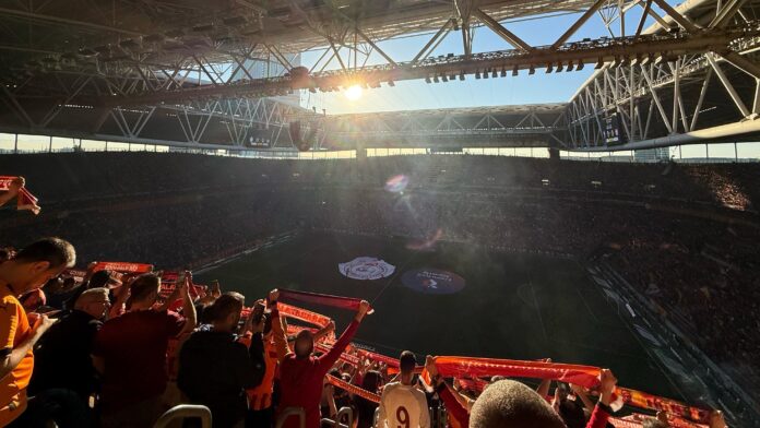 Wide view of Galatasaray fans holding scarves at Rams Park with sunlight streaming over the stadium during a match.