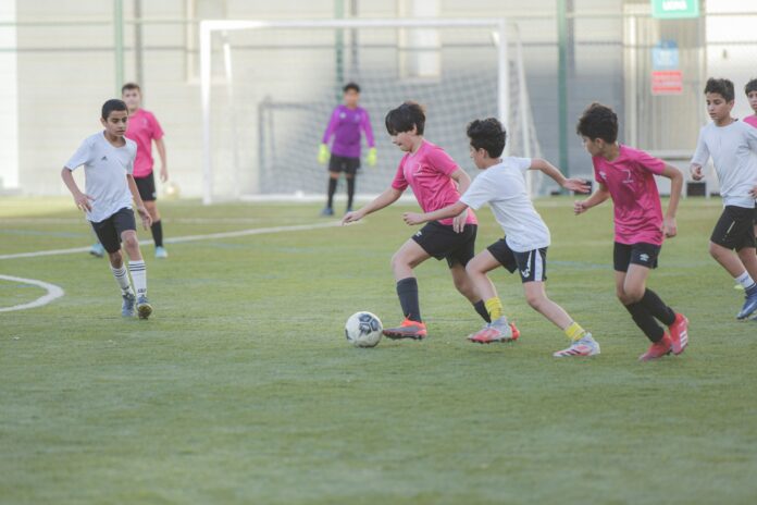 Youth football players in pink and white jerseys compete during a training match on a green pitch, with a goalkeeper in the background near the goal.