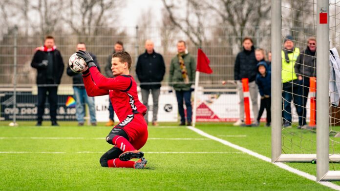 Young goalkeeper making a save during a senior football match in front of spectators.