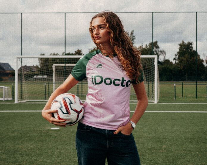 A woman holds a football on the pitch wearing Essevee Women’s pink and dark green 2026/27 away shirt.