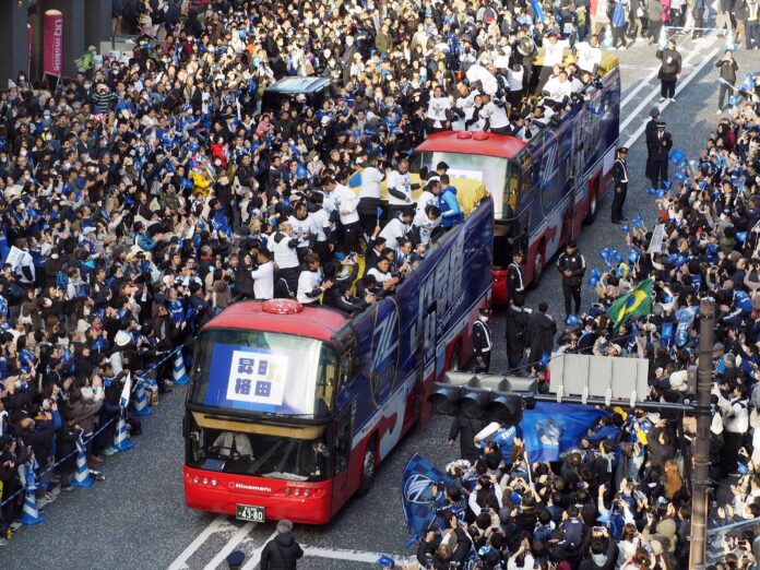 FC Machida Zelvia players celebrate their 2023 J2 title on open-top buses surrounded by supporters.