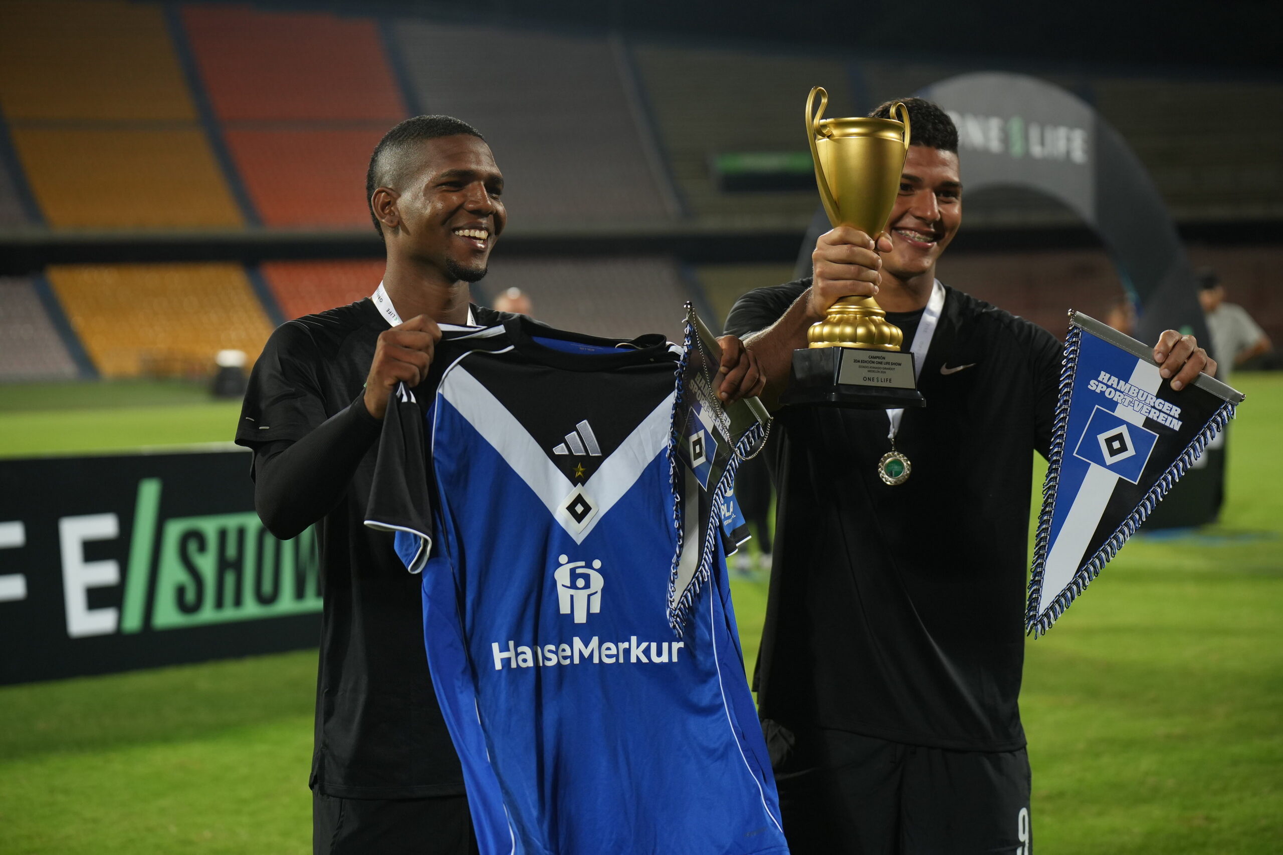 Two One-Life Show players celebrate with a trophy, Hamburger SV shirt and club pennants in Medellin.