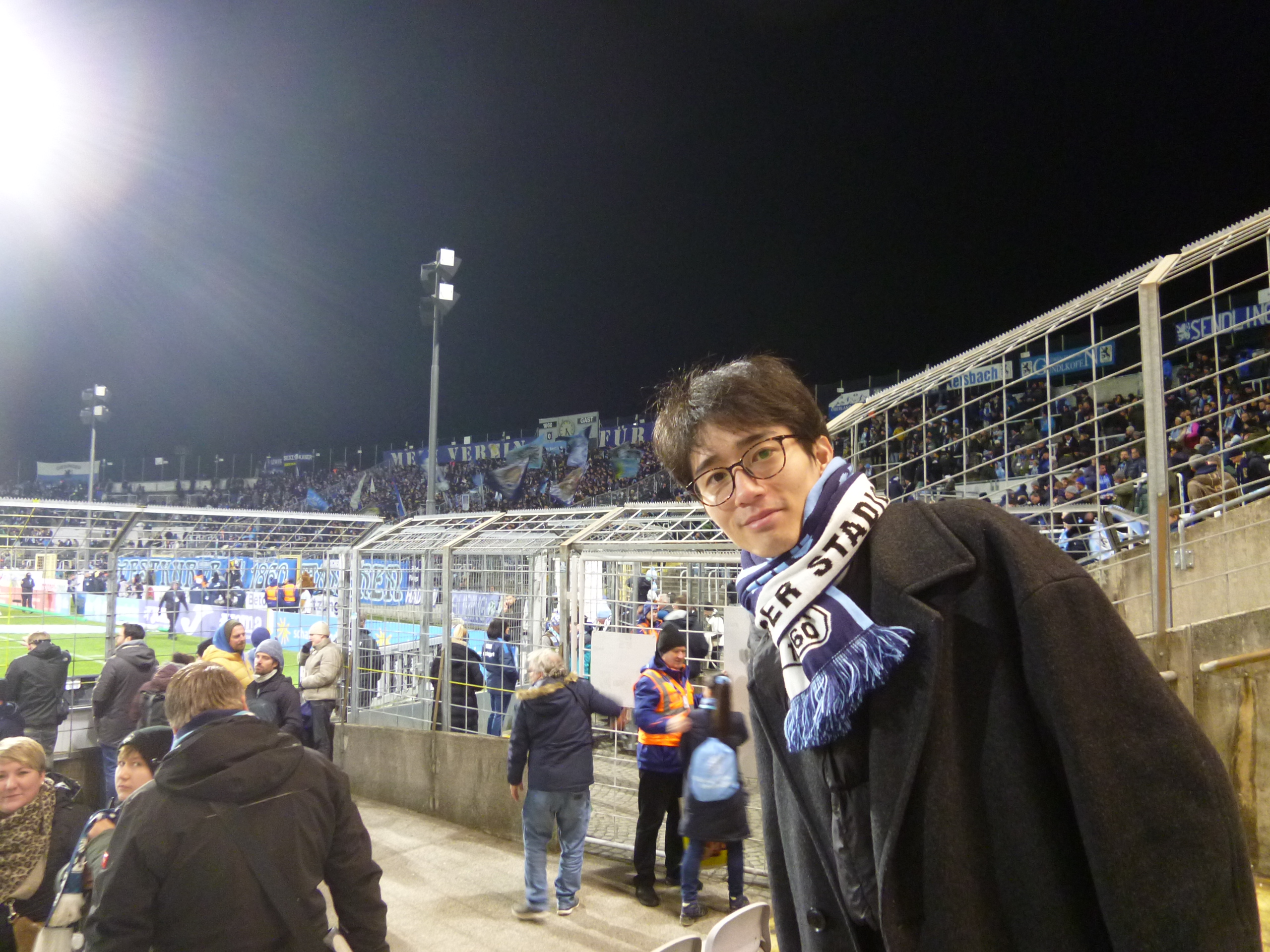 Ryusuke Hosaka wears a football scarf in the stands at a floodlit stadium.