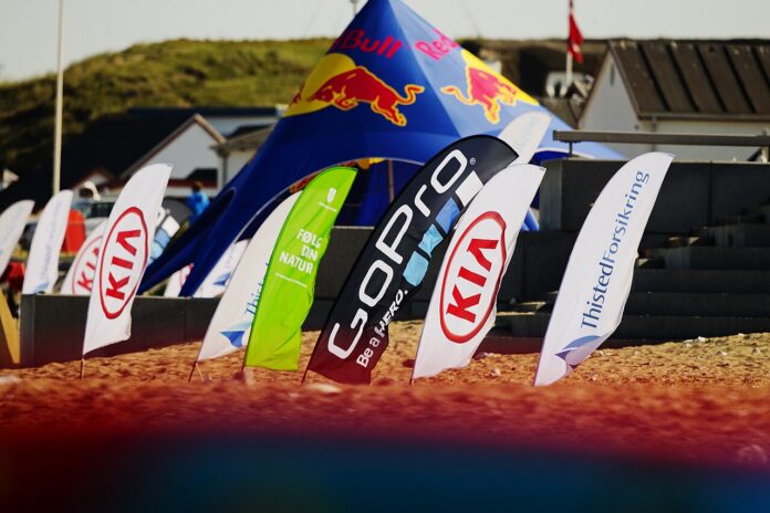 Sponsor flags from multiple brands line a beach sports venue during a commercial event.