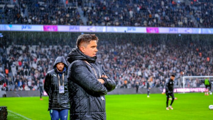 Bursaspor Chief Scout Batuhan Koça standing beside a football pitch in the rain with his arms folded.