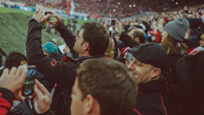 Football supporters capture the match on their smartphones from the stands of a crowded stadium.