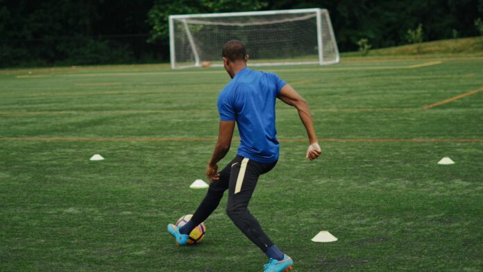 Football player in a blue shirt practising ball control among training cones on an outdoor pitch.